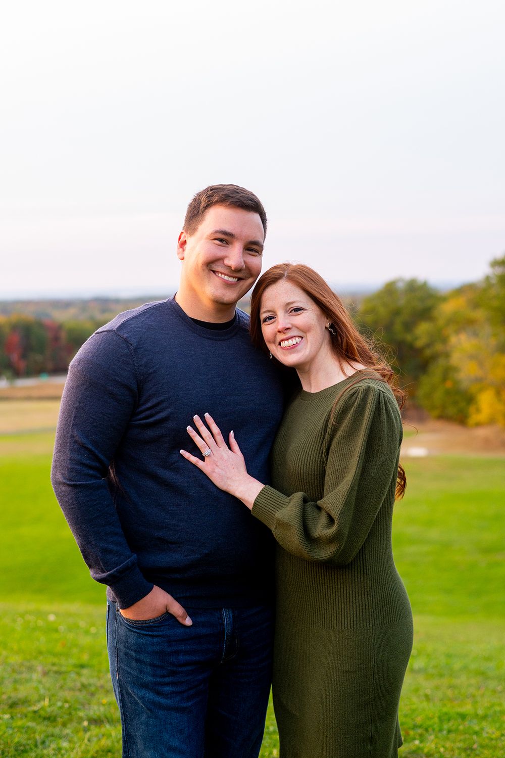 A joyful couple embraces in a grassy field during autumn while showing off an engagement ring.