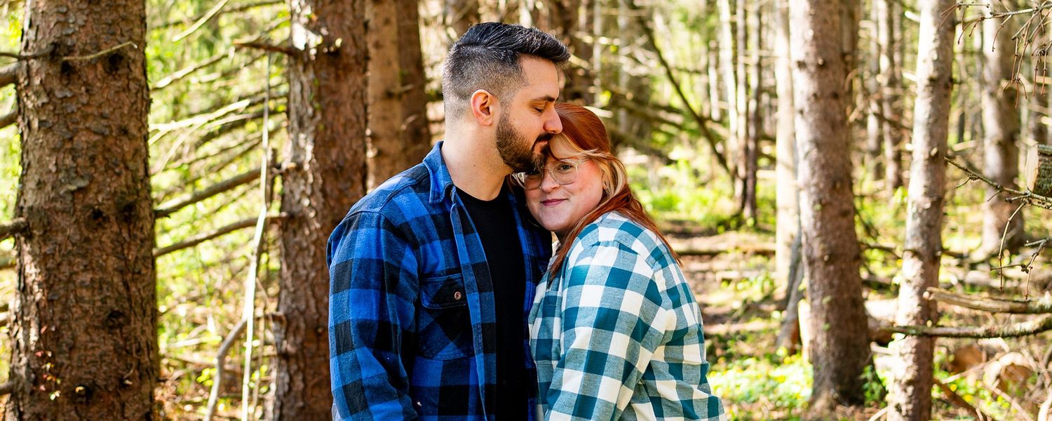 A couple in matching plaid flannel shirts embraces tenderly in a sunlit forest setting.
