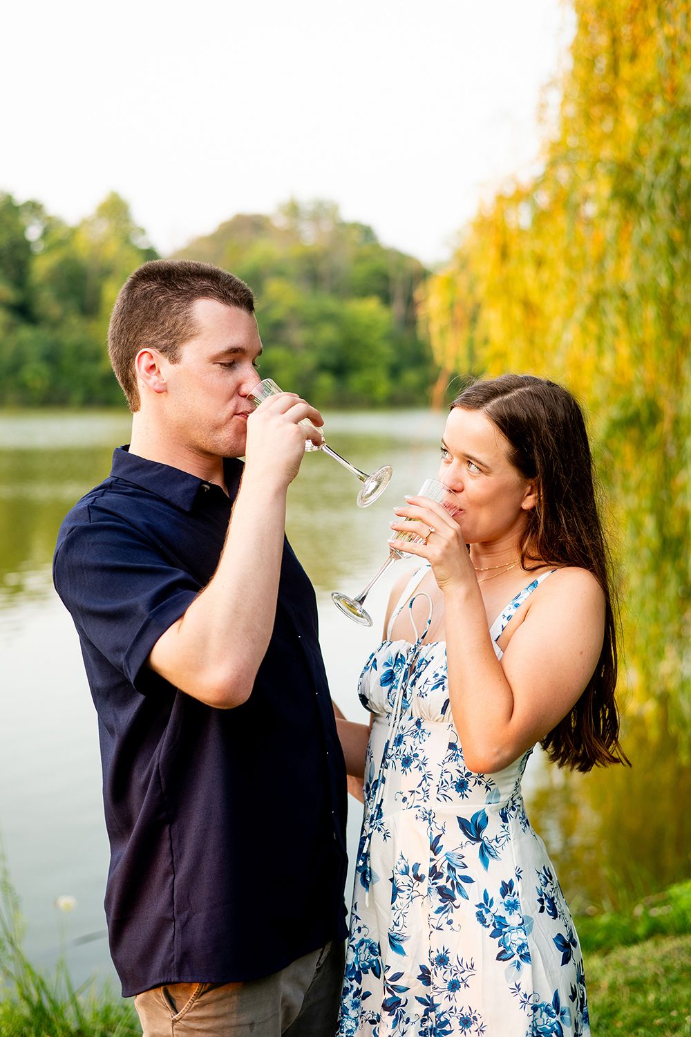 A couple shares a romantic moment with champagne glasses by a lake surrounded by autumn trees.