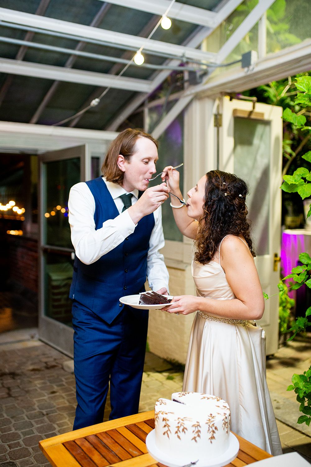 A couple shares wedding cake during their reception at a venue with glass ceiling and string lights overhead.