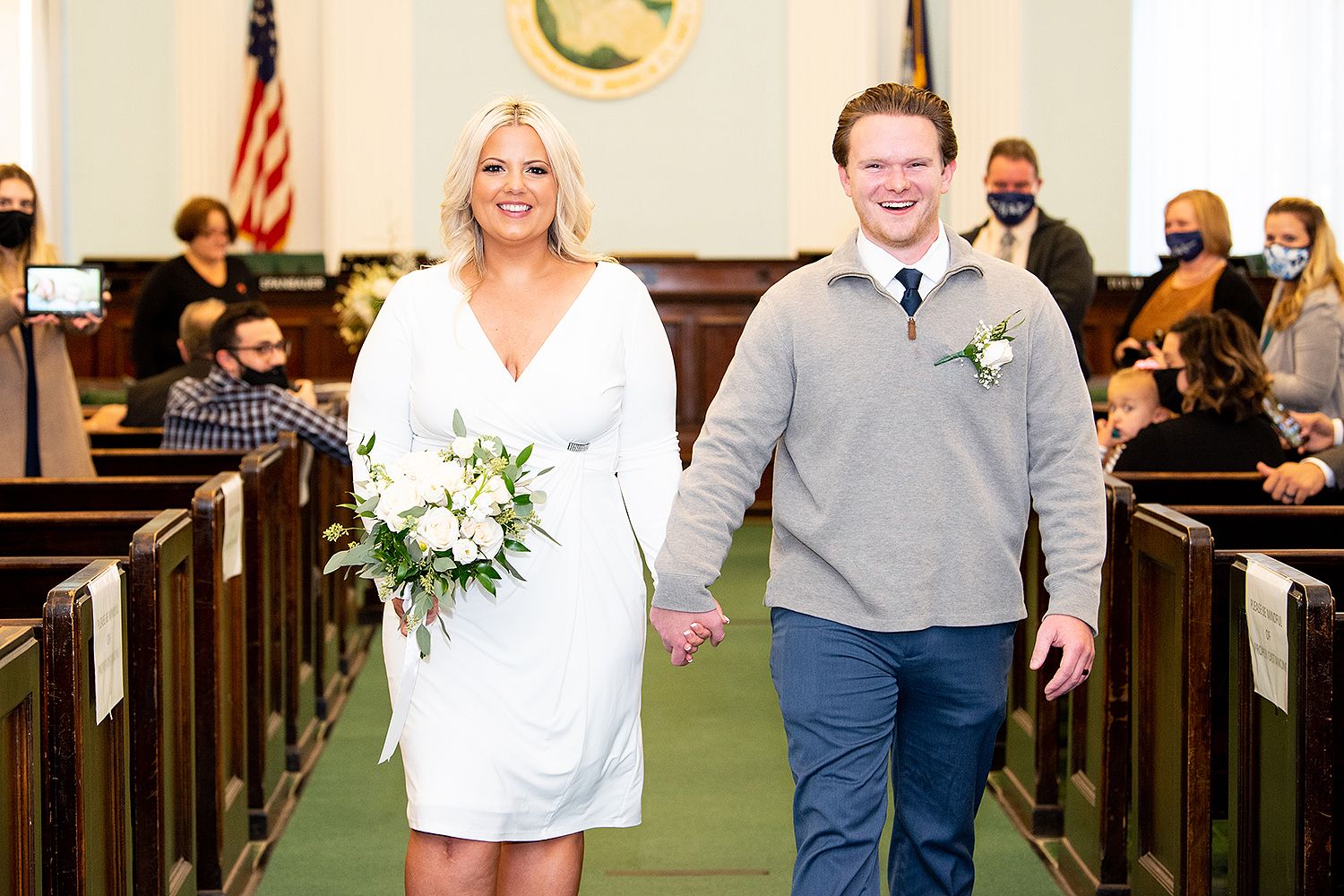Elegant wedding ceremony scene with wooden church pews, white floral arrangements and smiling people in formal attire.