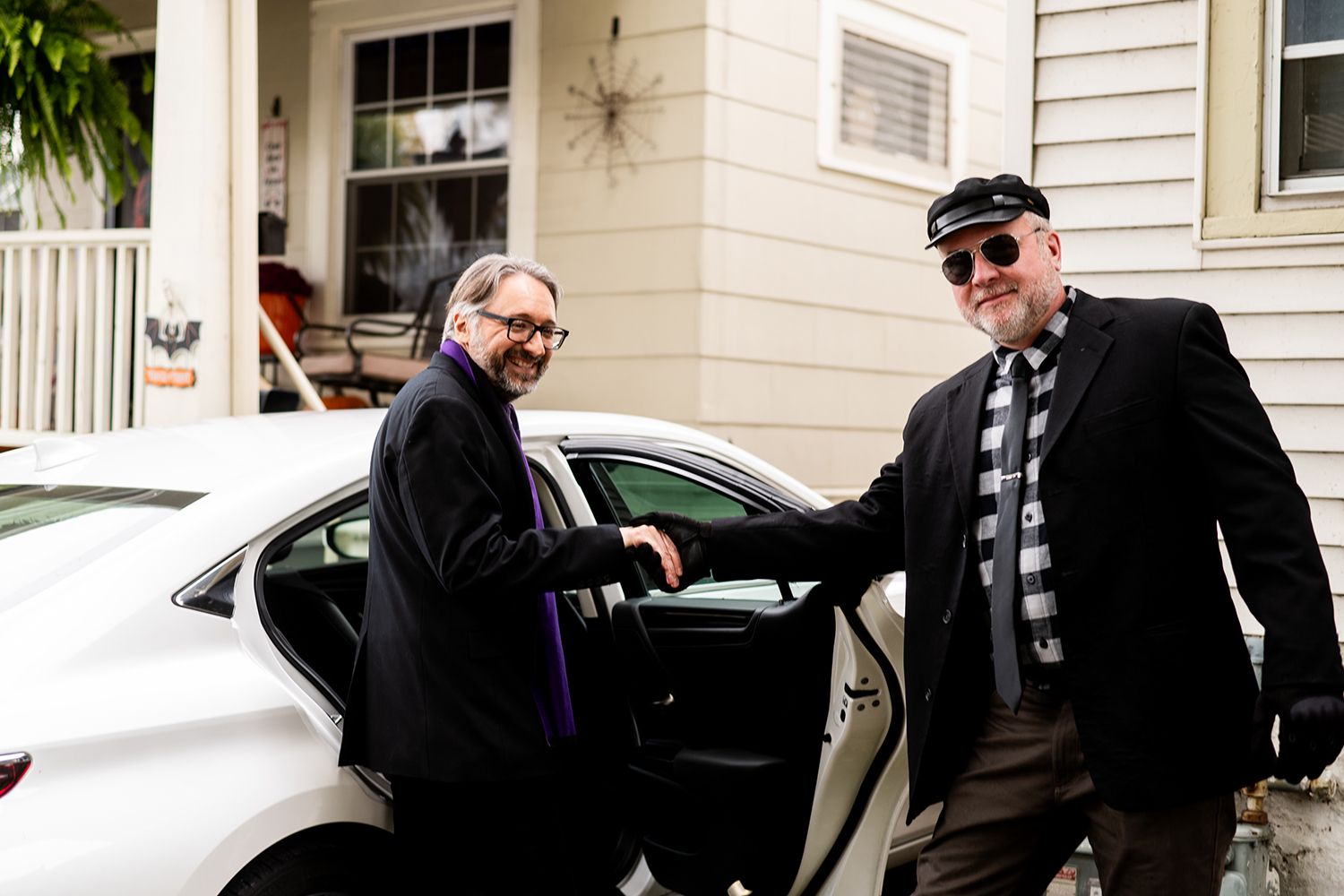 Two people share a laugh while standing next to a white car during a wedding event.