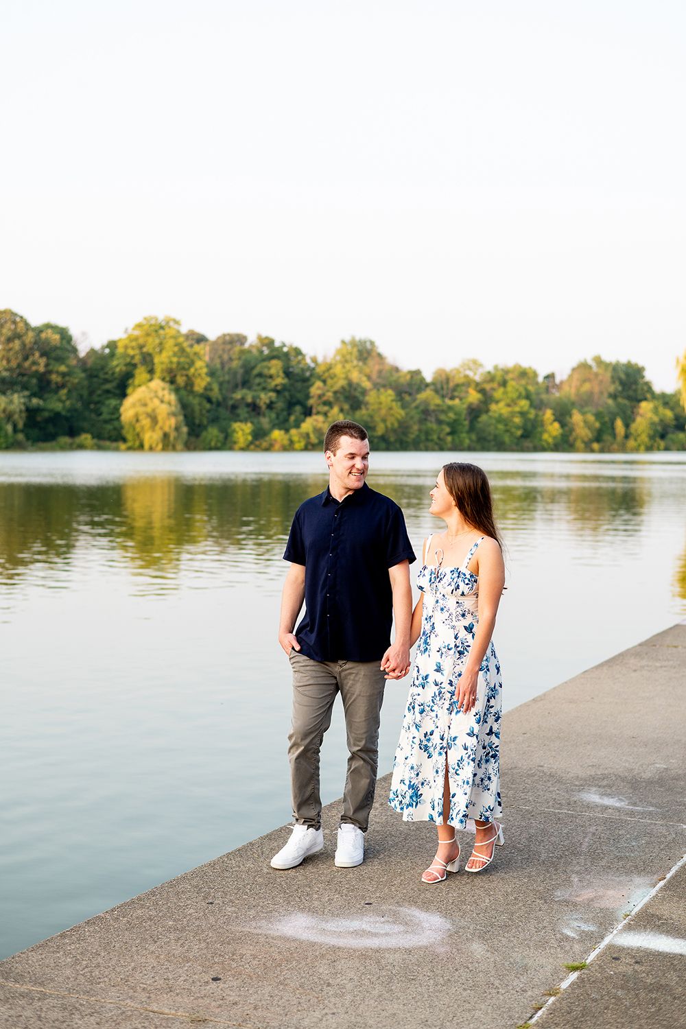Young couple holding hands while walking along lakeside path during golden hour with fall foliage reflecting in calm water.
