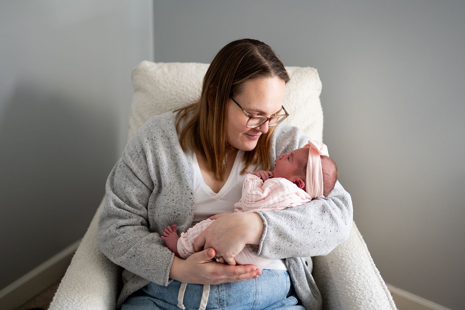 A mother sits in a white armchair cradling her newborn baby in a soft blanket.