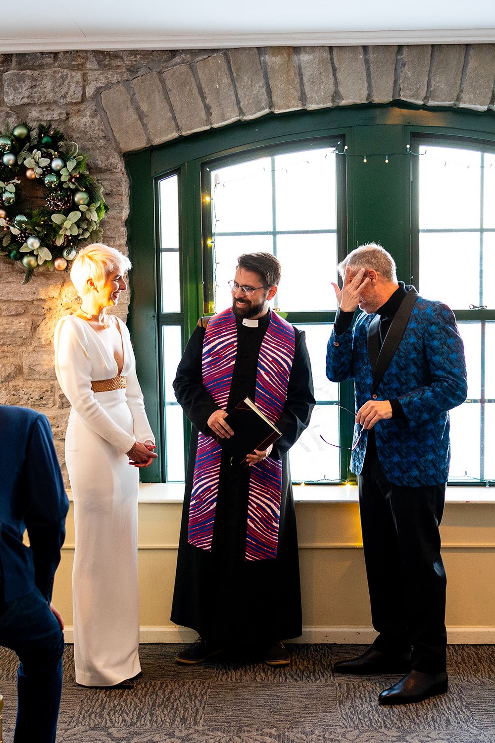 Wedding ceremony taking place in historic stone room with arched windows and clergy member wearing purple stole.