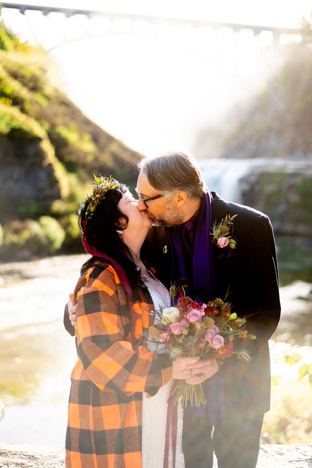 A couple sharing a romantic kiss at sunset by a waterfall with a bouquet of pink flowers and golden sunlight.
