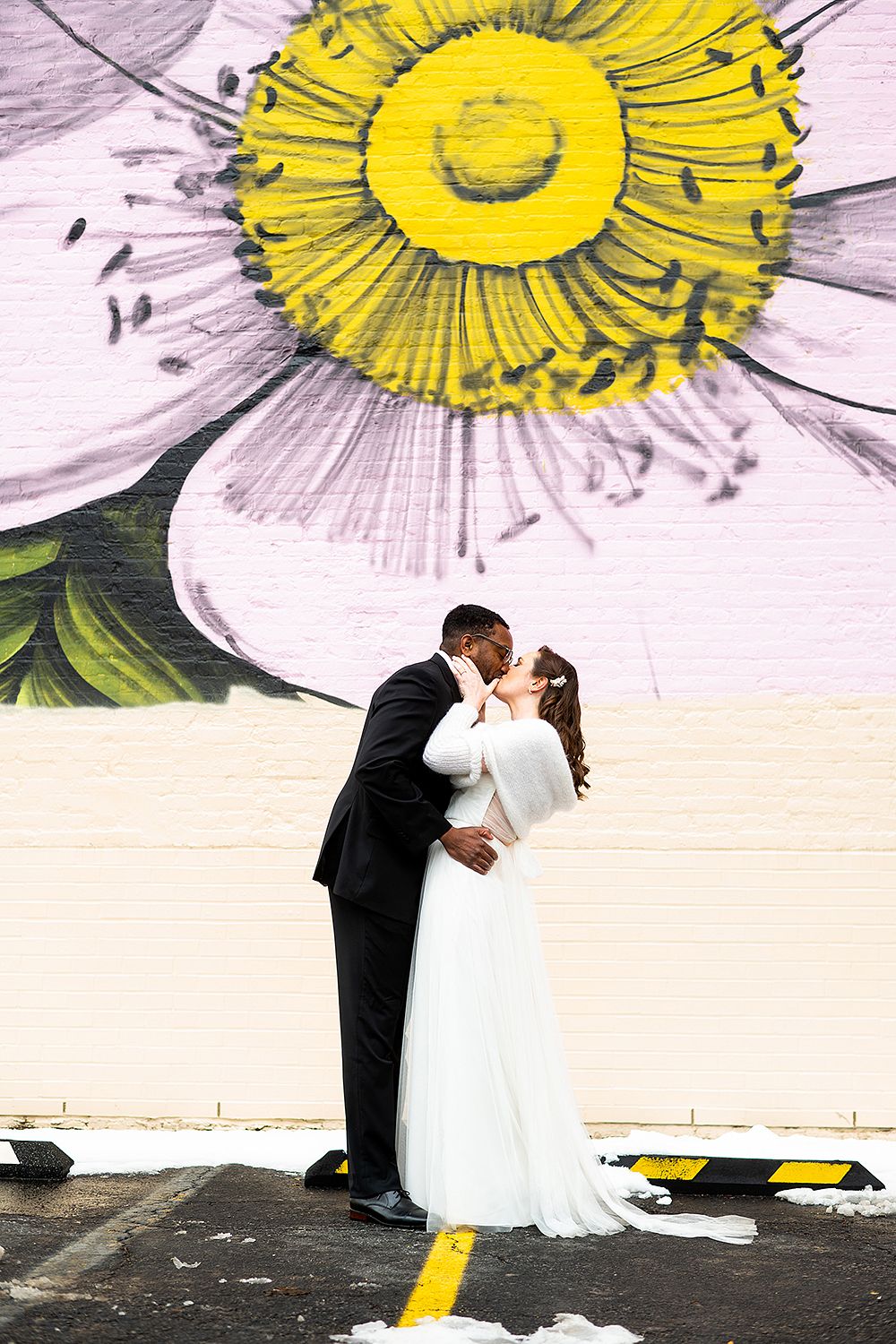 A couple embraces in front of a large floral mural featuring white petals and a bright yellow center.