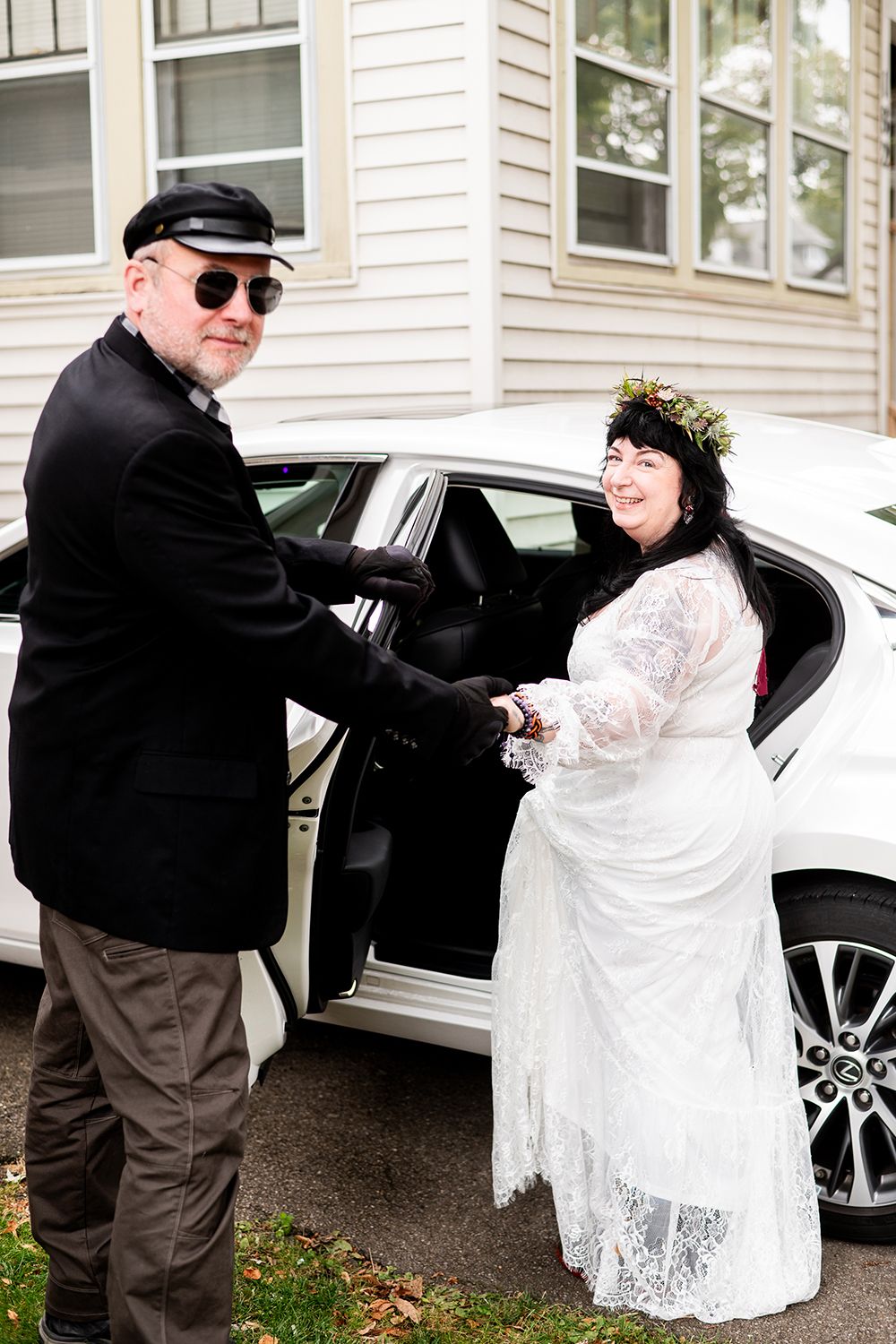 A wedding photo of a couple sharing a joyful moment by a white car in front of a house.
