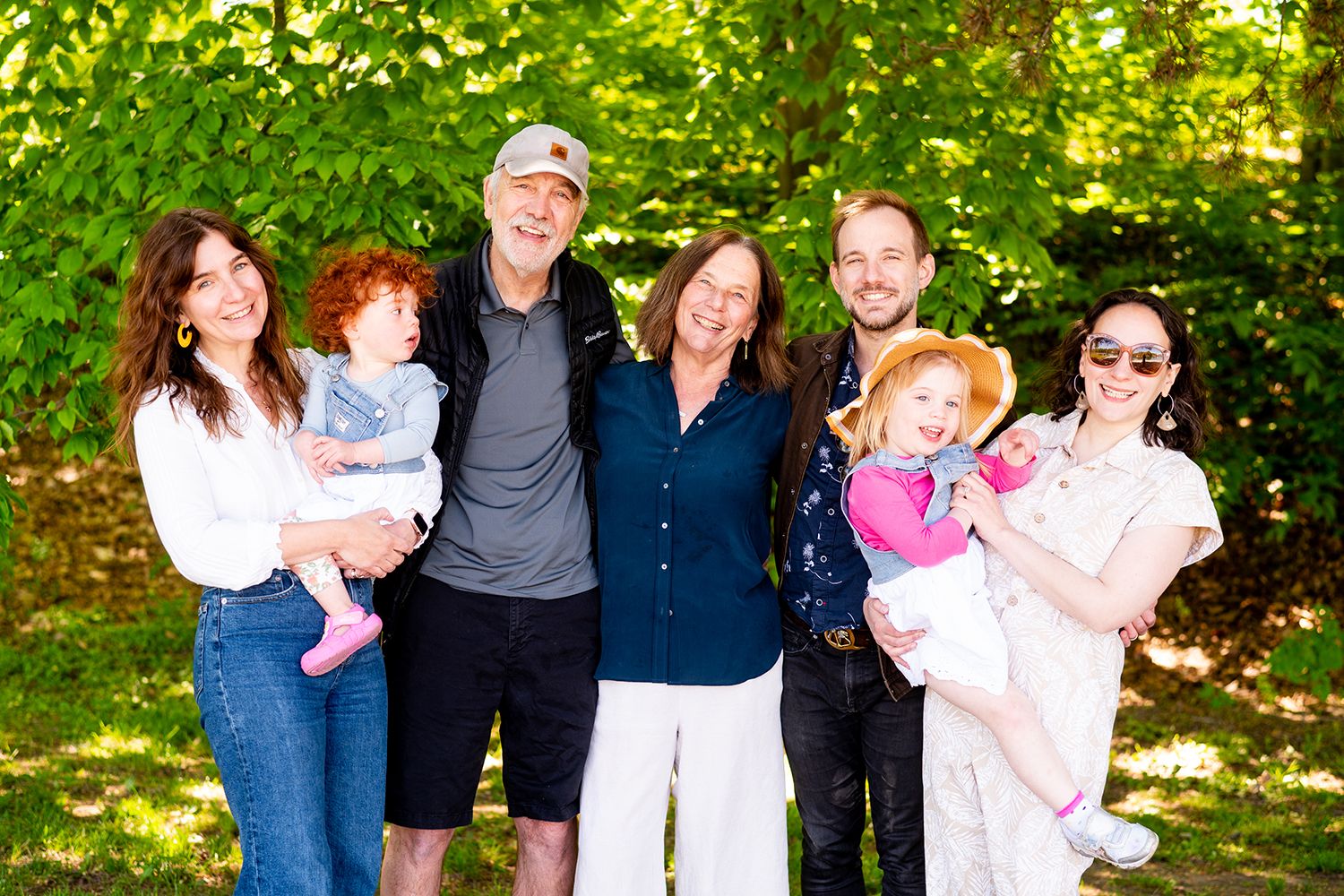 Multi-generational family group photo smiling together outdoors with lush green trees in background.