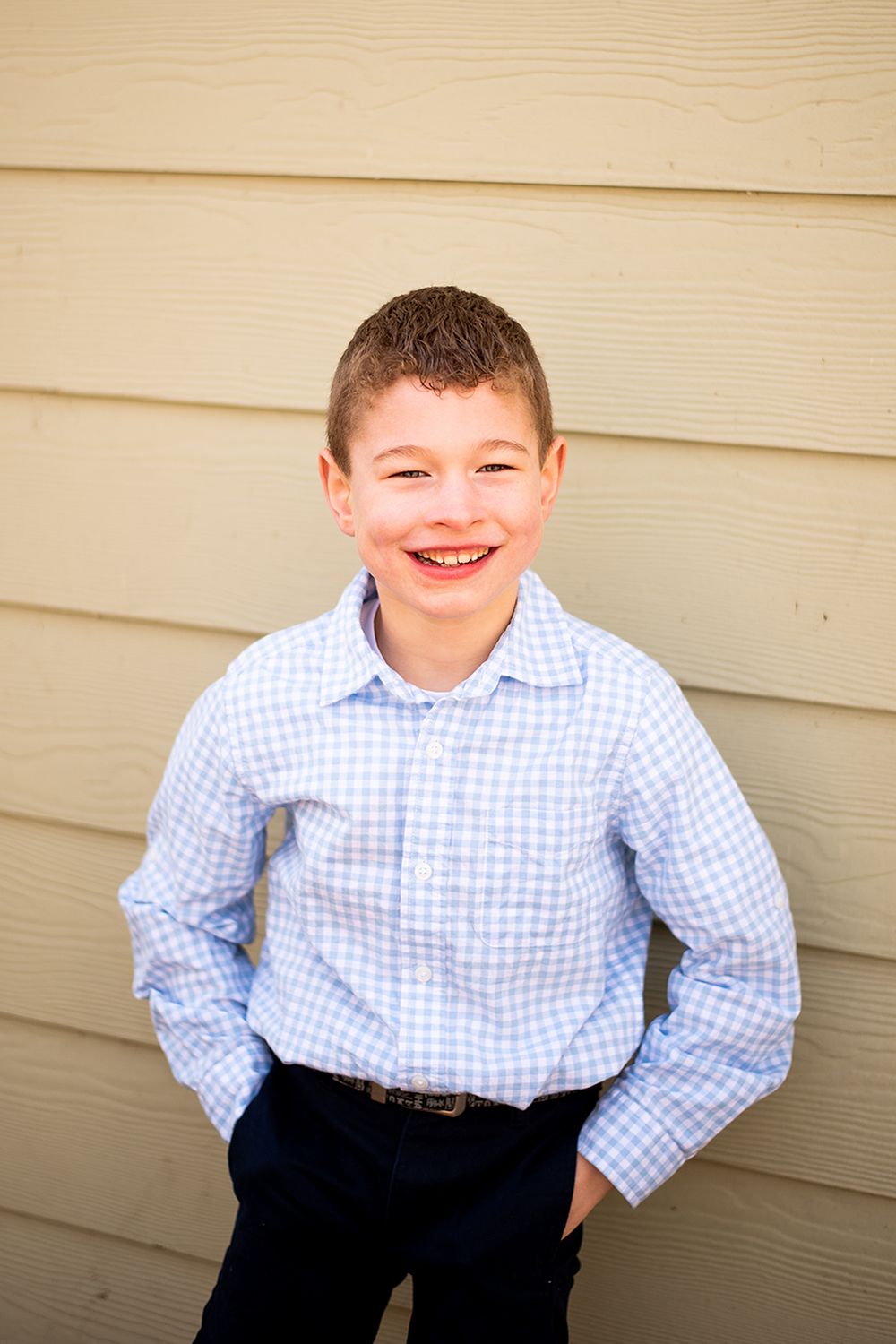 Cheerful portrait against tan siding wearing light blue checkered button-up shirt.