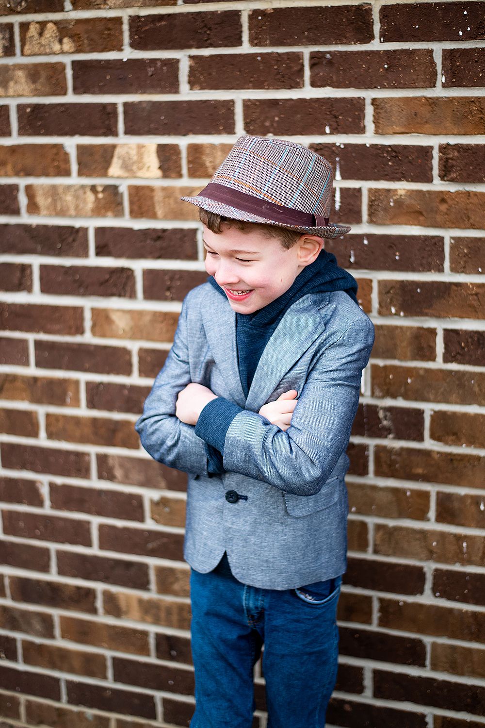 Stylishly dressed in a gray blazer and plaid hat against a brick wall with crossed arms and joyful expression.