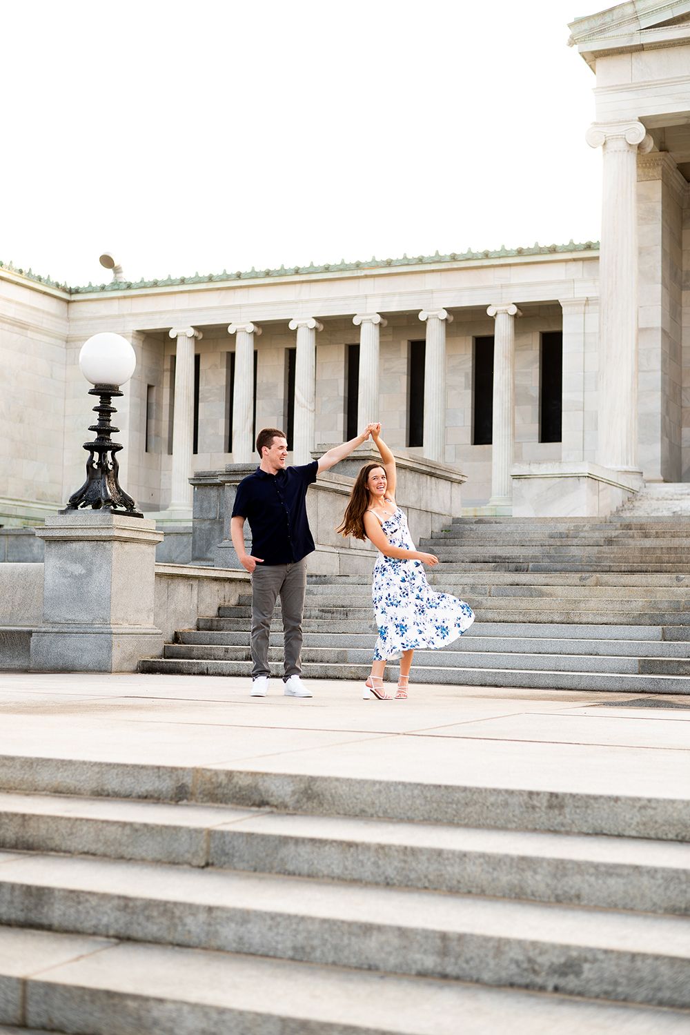 Two people dance on the grand stone steps of a neoclassical building with elegant white columns and globe lamps.