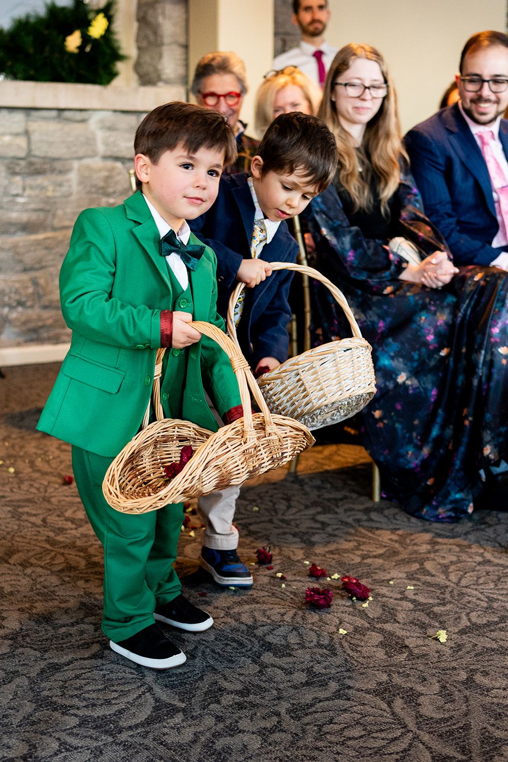 A young attendant in a green jacket tosses flower petals from a woven basket during an outdoor wedding ceremony.