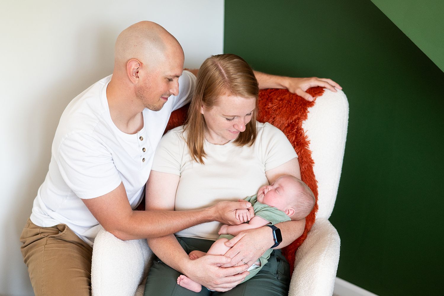 A family sits together in a white armchair, sharing a tender moment while holding a sleeping baby.