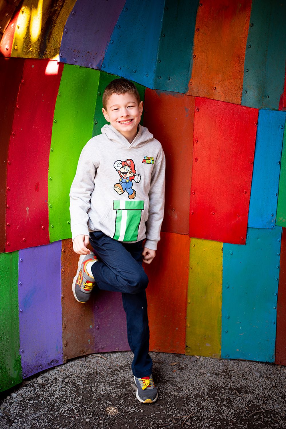 Super Mario fan poses playfully against vibrant rainbow-colored wall in gray Nintendo sweatshirt.