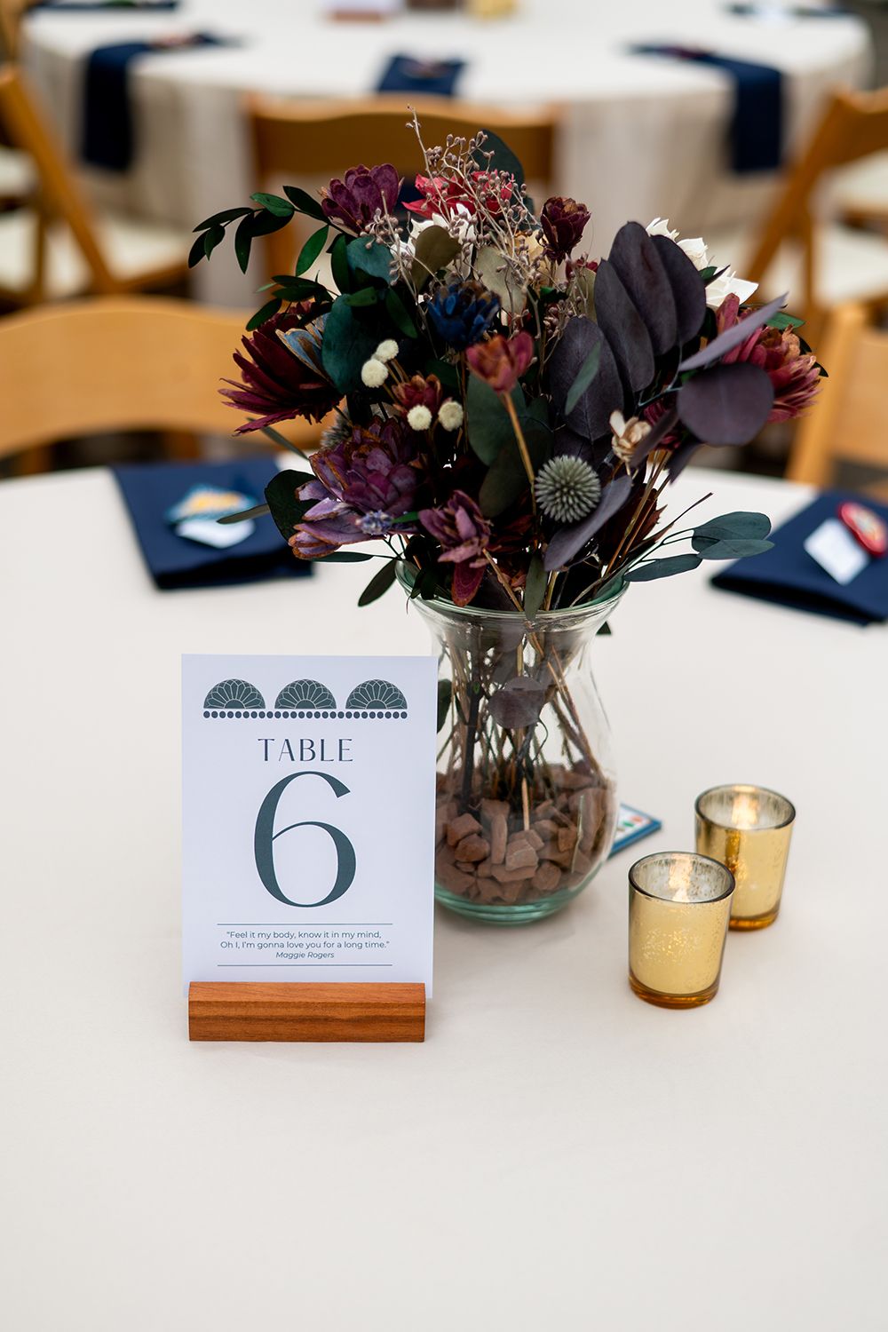 Dark moody floral centerpiece with table number 6 and gold votives on white tablecloth.
