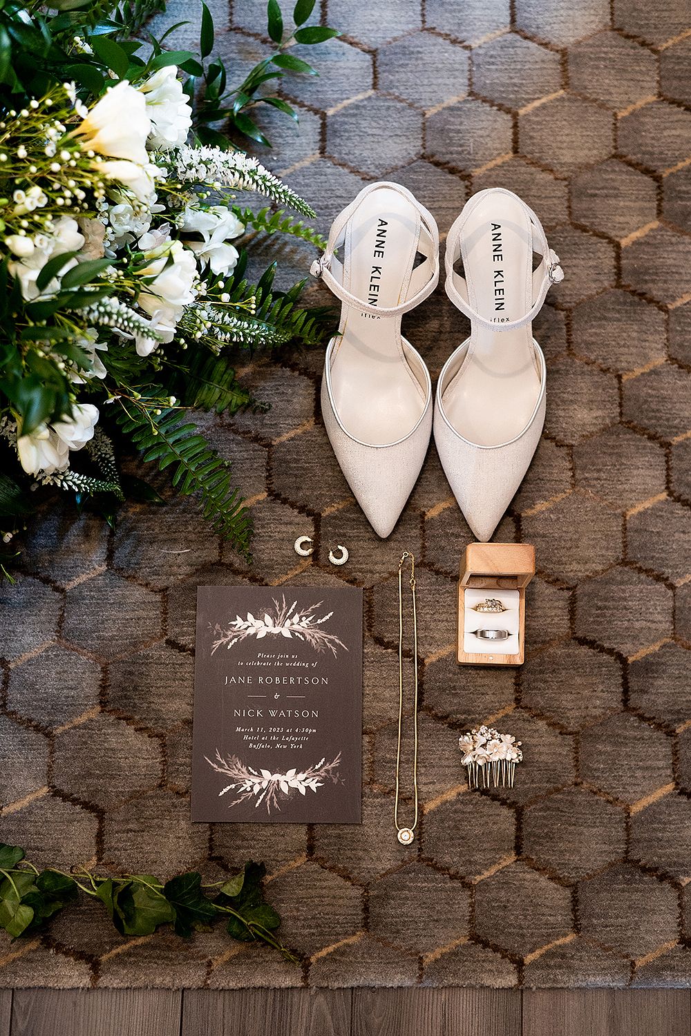 Wedding details flatlay with white heels, floral bouquet, dark invitation card and jewelry on textured gray background.
