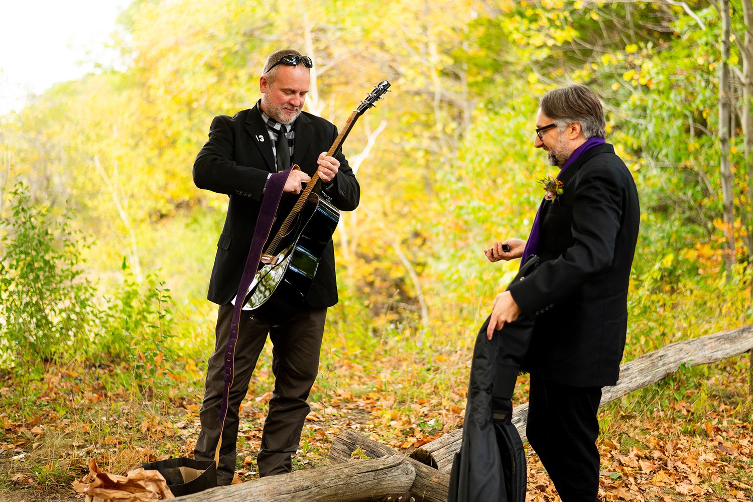 Two musicians in black suits perform outdoors during autumn with stringed instruments in a wooded setting.