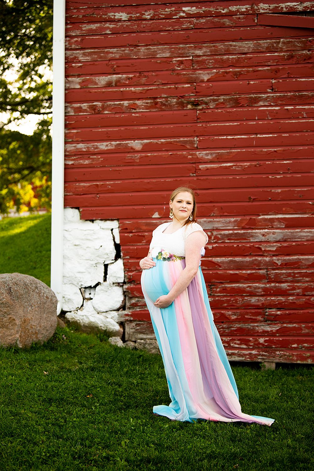 Elegant maternity photo taken against a rustic red barn with white stone foundation on a sunny summer evening.