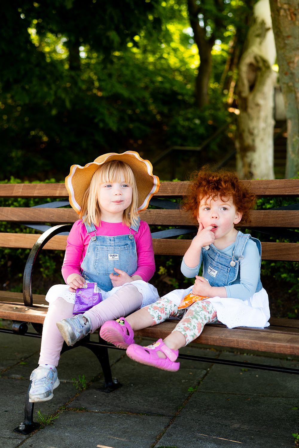 Two children in overalls sitting on a park bench, one wearing a sun hat and the other making silly expressions.