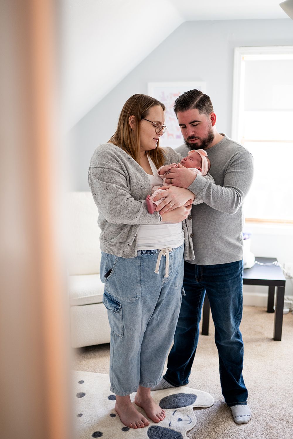 A couple stands together holding their newborn baby in a bright room with white walls and carpeted floor.