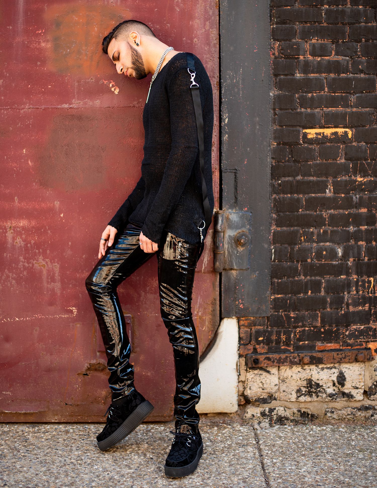 Person in black sweater and metallic striped pants leaning against rustic brick wall with industrial door.