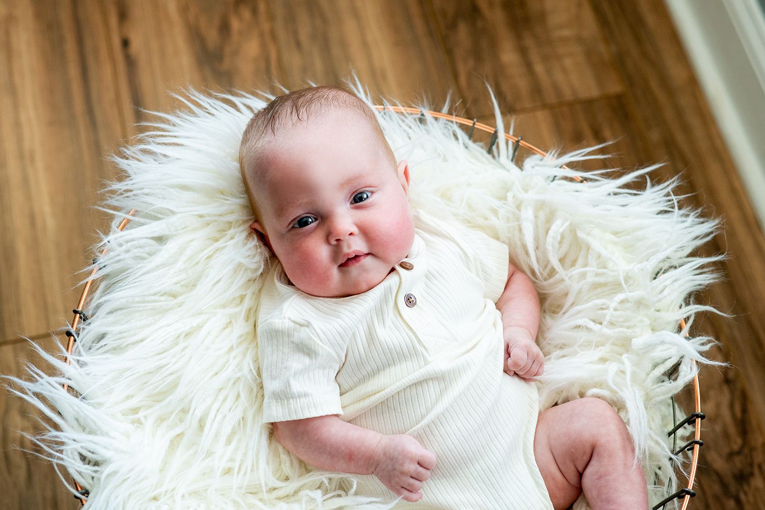 Baby lying on soft white fur rug wearing white onesie on wooden floor.