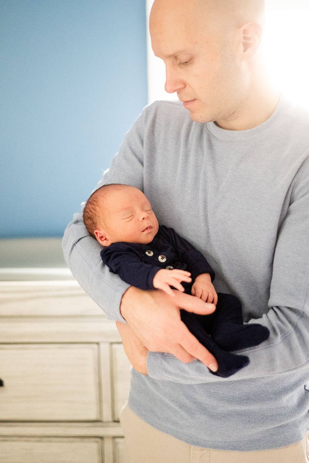 A tender moment shows a person in a gray sweater cradling a sleeping newborn baby wearing a navy outfit.