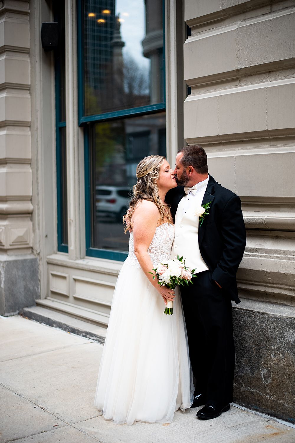 Bride in white wedding gown and groom in black suit share intimate moment against stone building exterior.