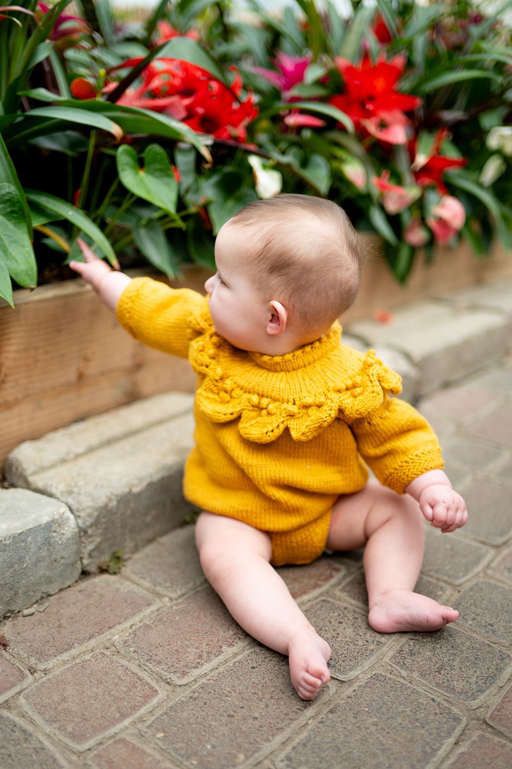 Baby wearing knitted yellow sweater crawls on brick patio near flowering plants.