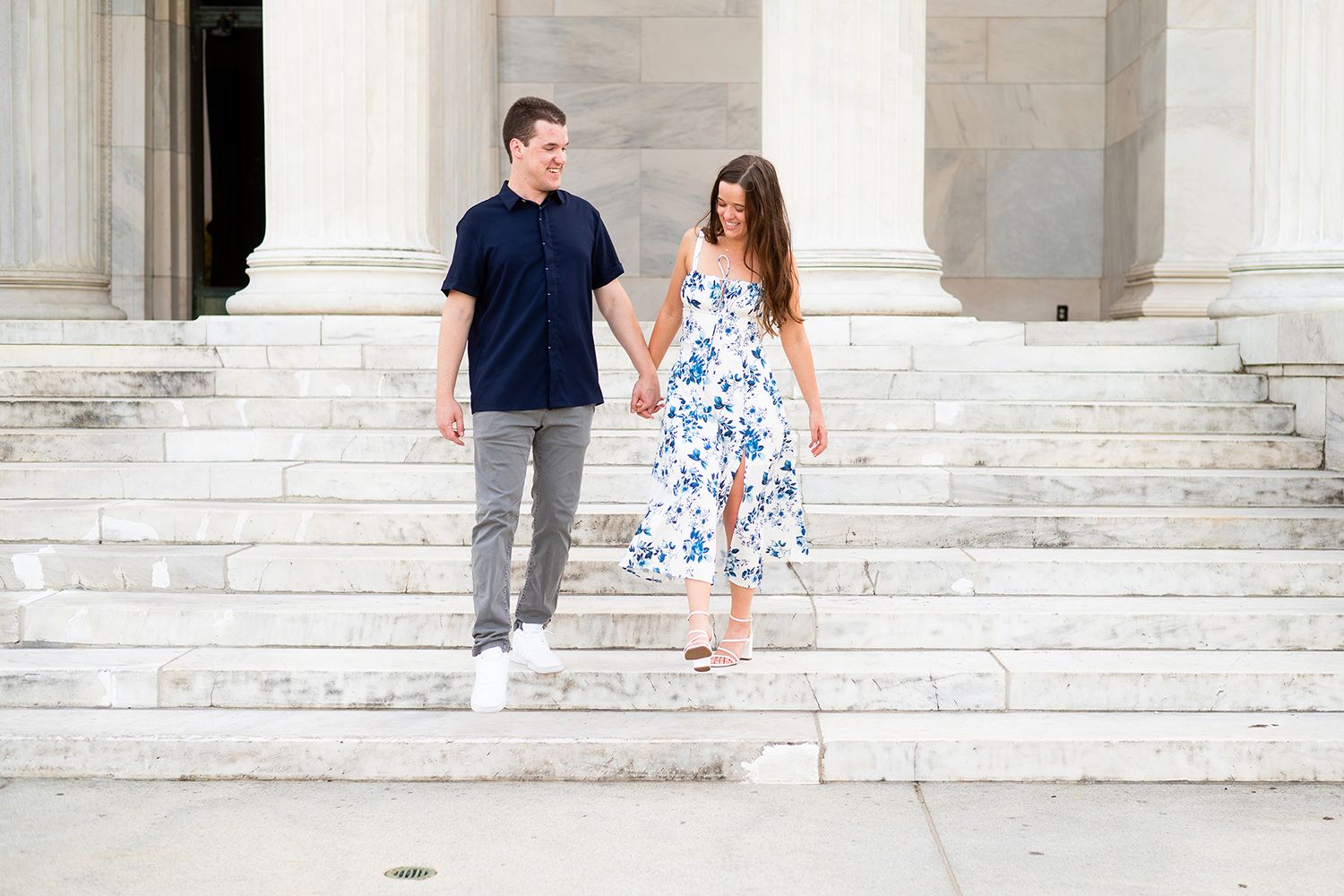 A couple walks down white marble steps at a grand historic building, with the woman wearing a floral summer dress.