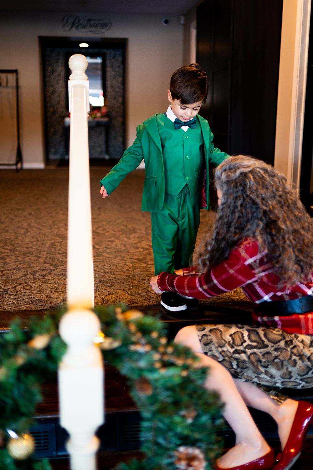 A child in a bright green outfit stands near holiday decorations and a tall white candle in an elegant hallway.