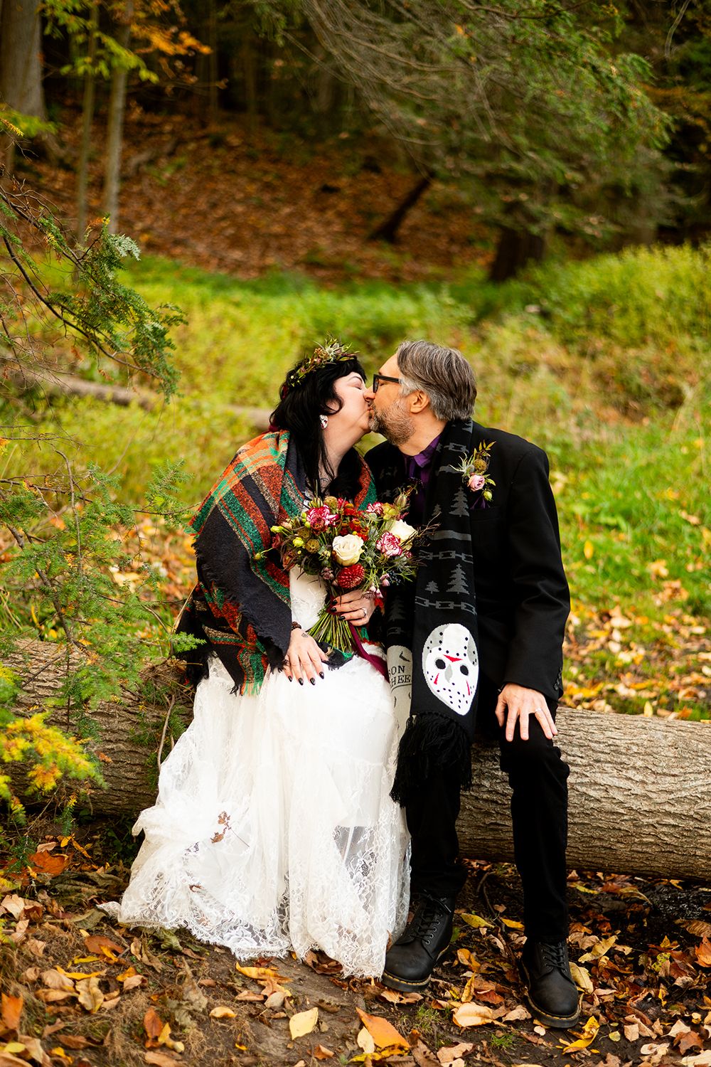 A couple in Halloween wedding attire share a romantic moment on a fallen log surrounded by autumn foliage.