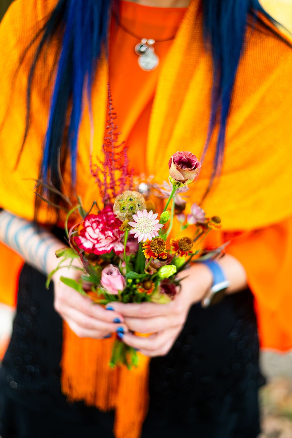 Person wearing bright orange top and blue hair holds small colorful floral bouquet featuring pink blooms and greenery.