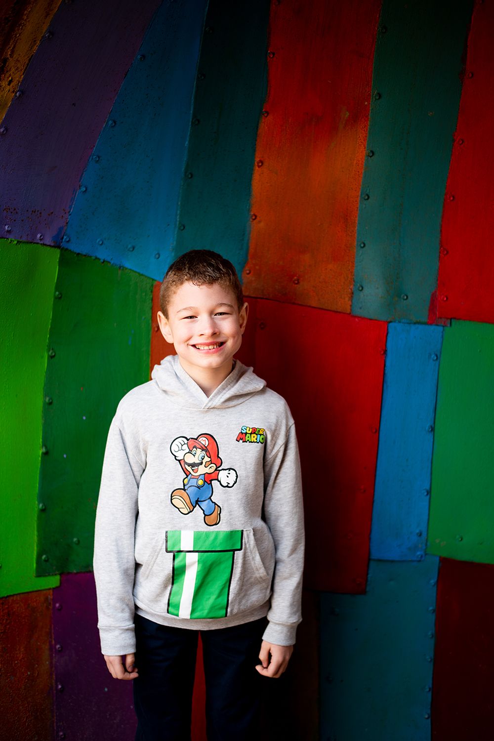 Young person wearing gray Super Mario hoodie smiles against colorful striped wall background.