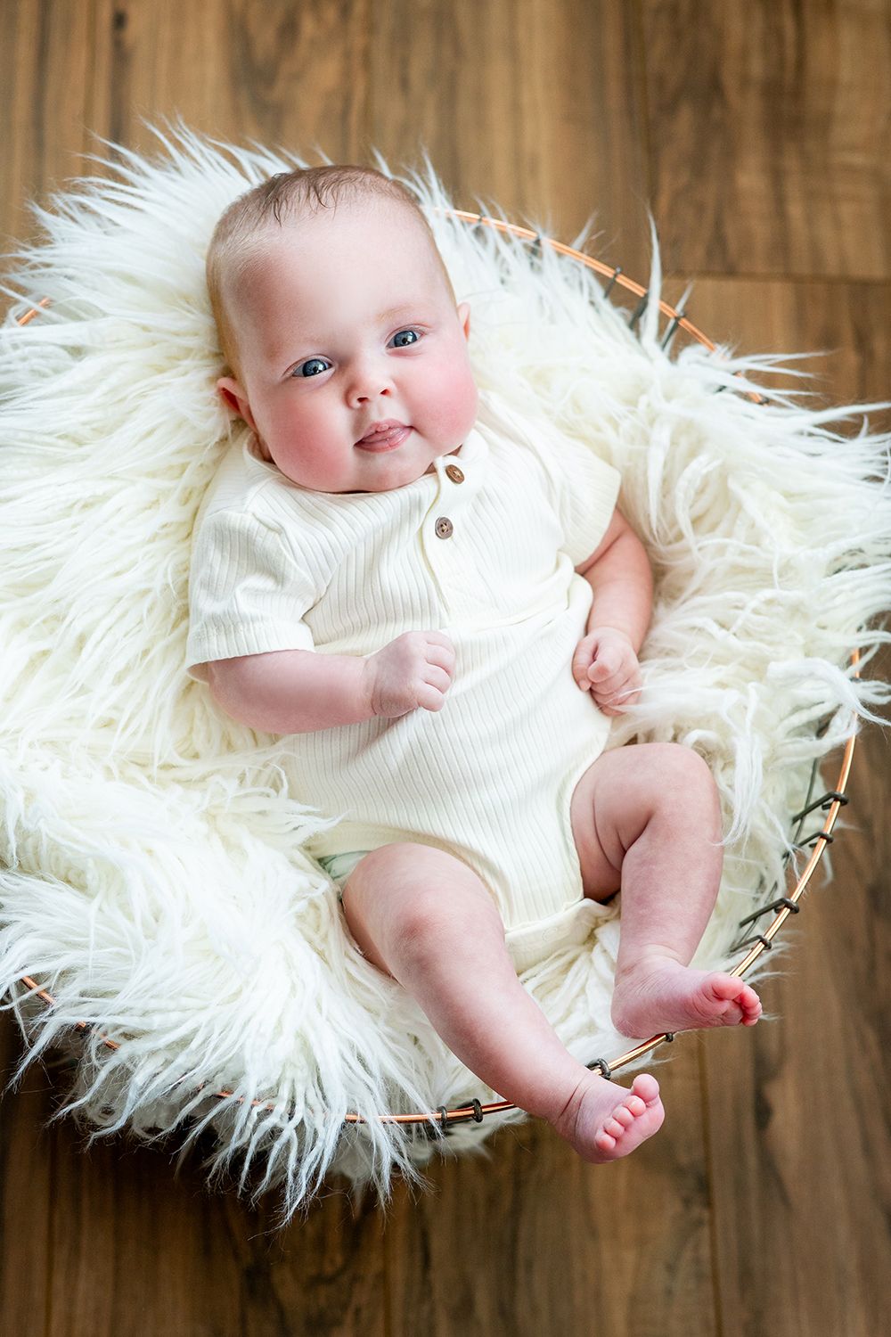 A baby in a white onesie lies on a plush faux fur white rug against a wooden floor background.
