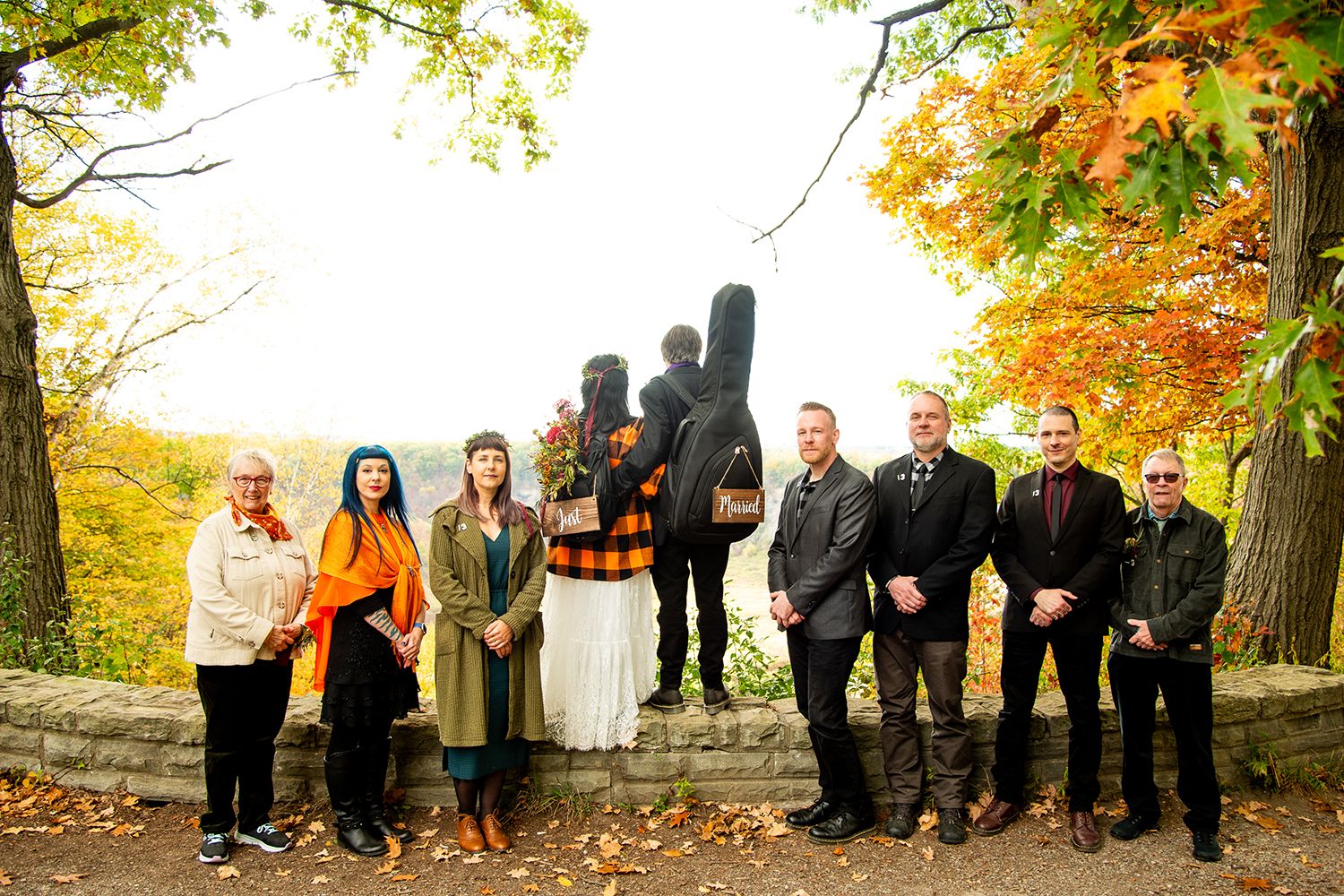 Wedding party poses for group photo with autumn foliage and silhouetted sculpture in background.