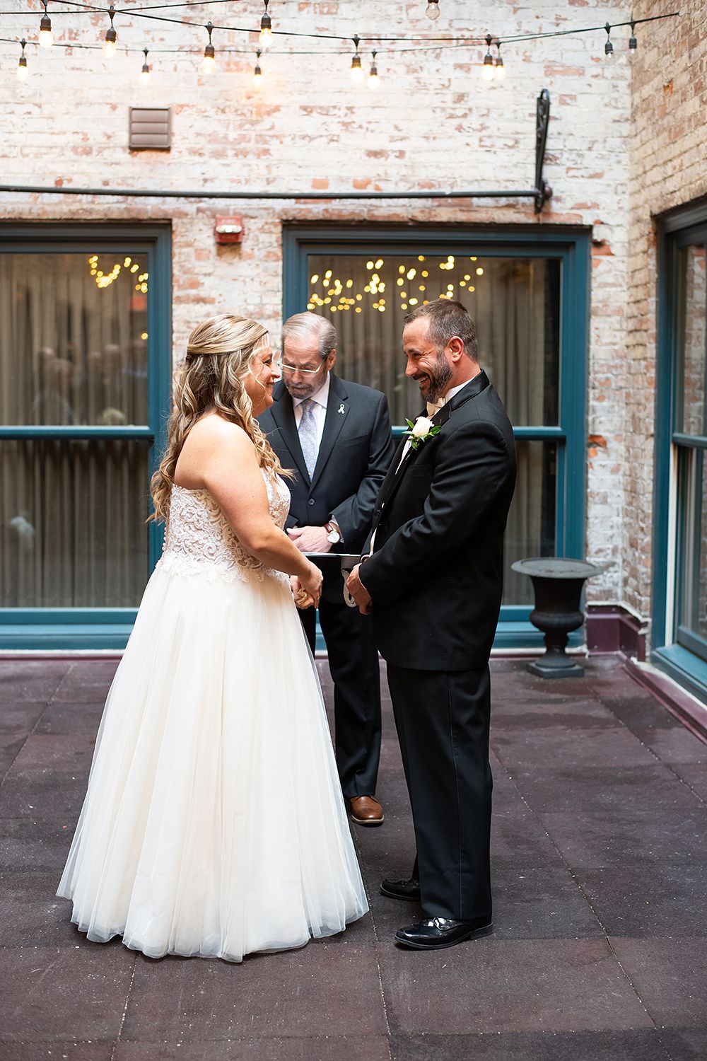 A wedding ceremony takes place in a courtyard with exposed brick walls and string lights overhead.