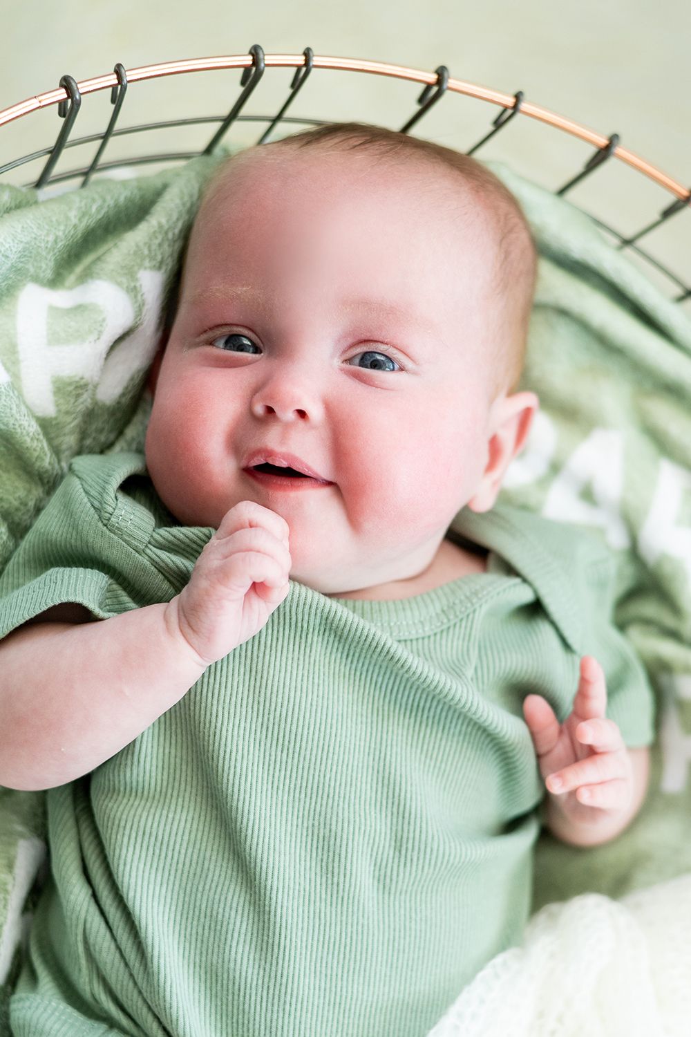 A happy baby wearing a mint green outfit lies on soft bedding while sucking their thumb.