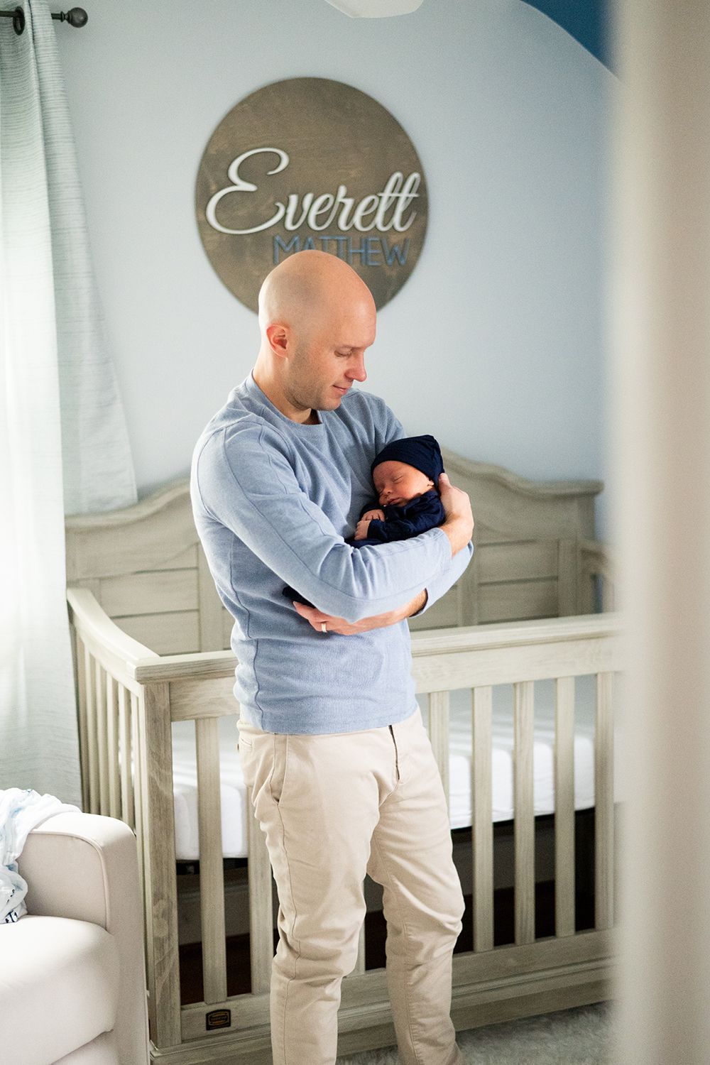 A parent wearing a gray sweater holds a newborn baby in a nursery room with the name Everett displayed on the wall.