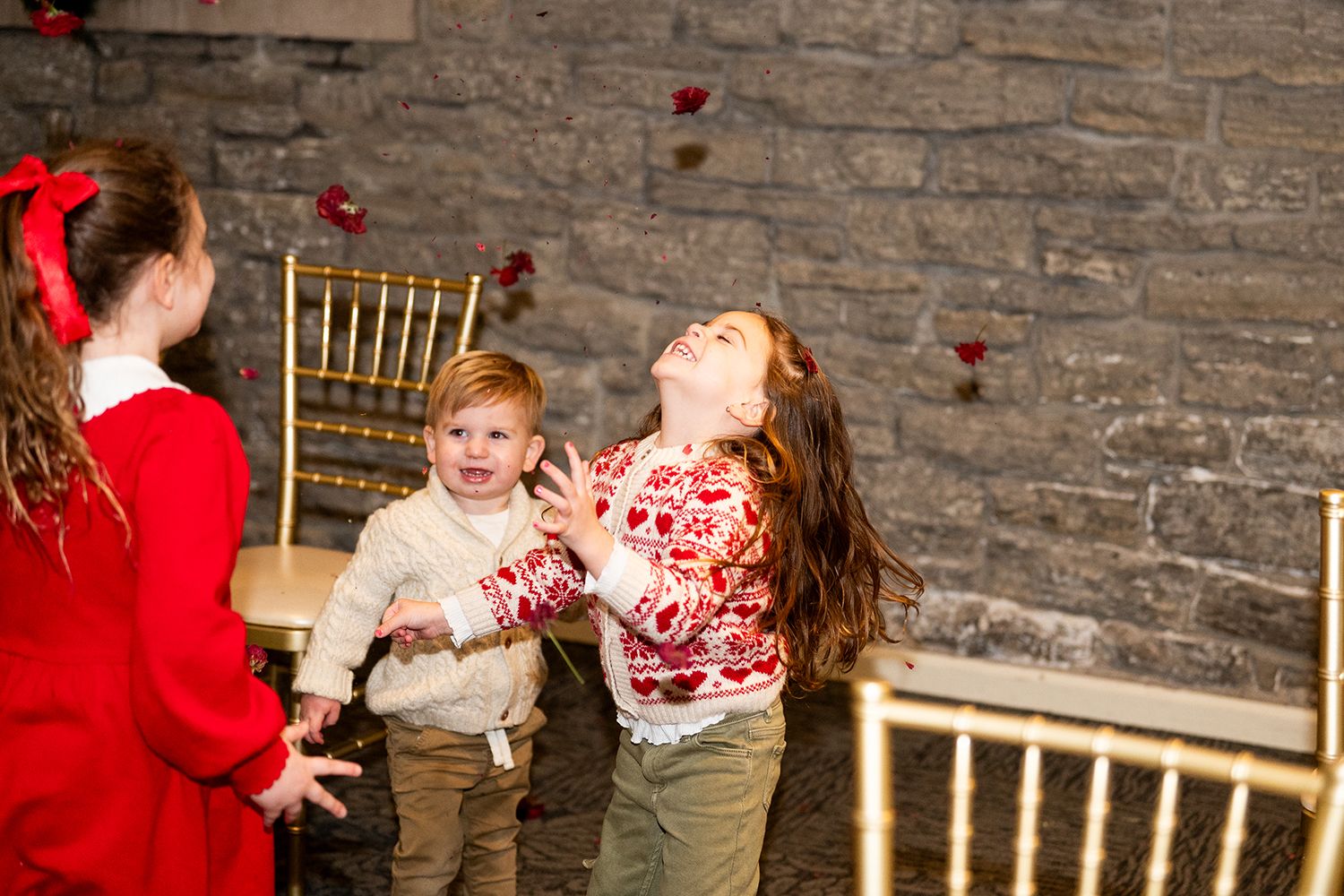 Children laughing and playing with red petals in front of a rustic stone wall with gold chairs.