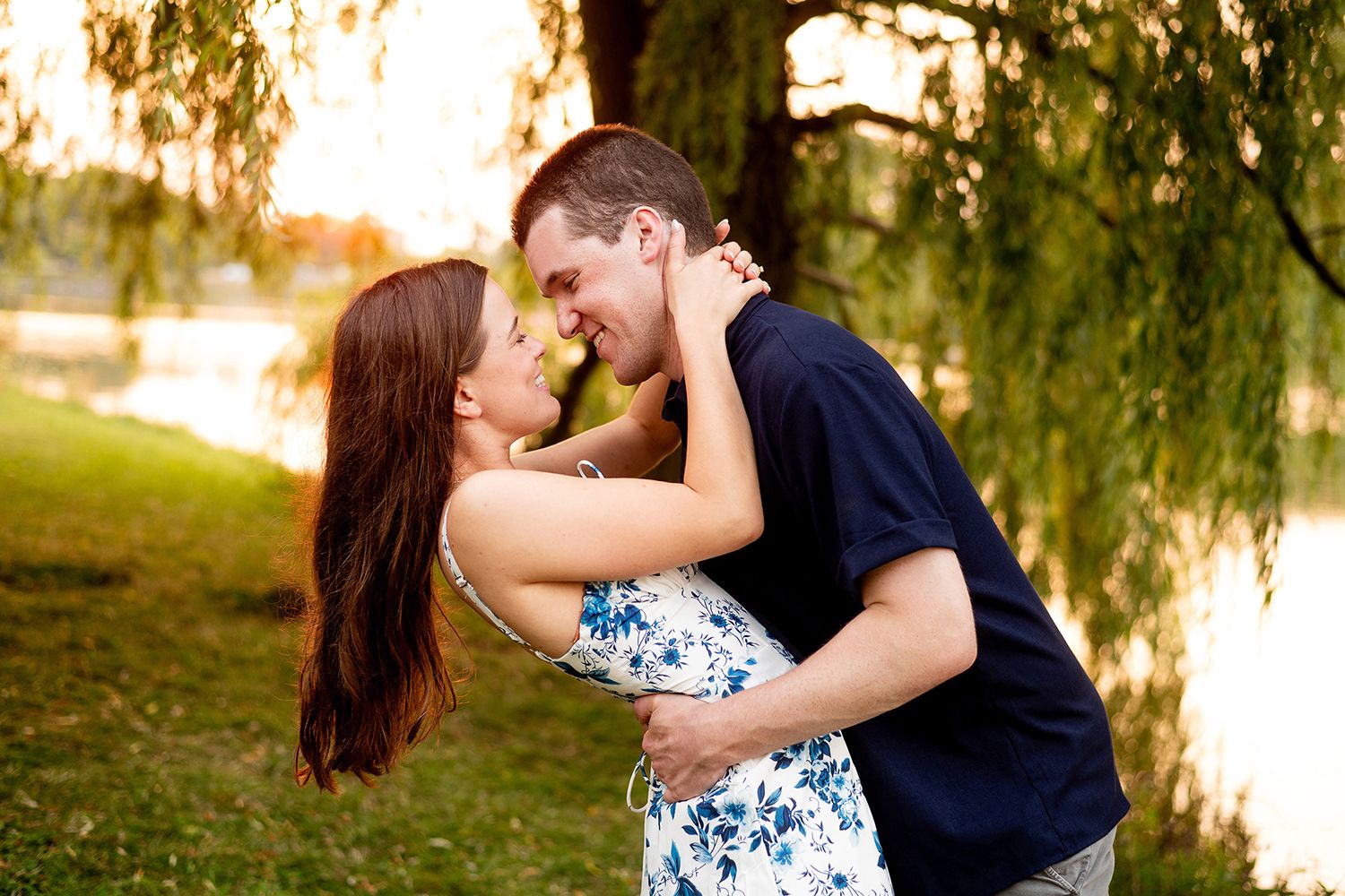 A romantic couple embraces beneath drooping willow tree branches during a sunset photoshoot.