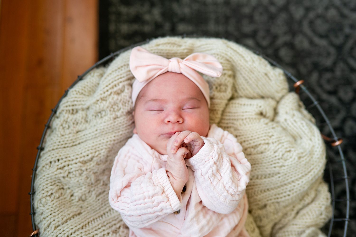 A sleeping baby swaddled in a pink bow outfit rests peacefully on a cream-colored knit blanket.