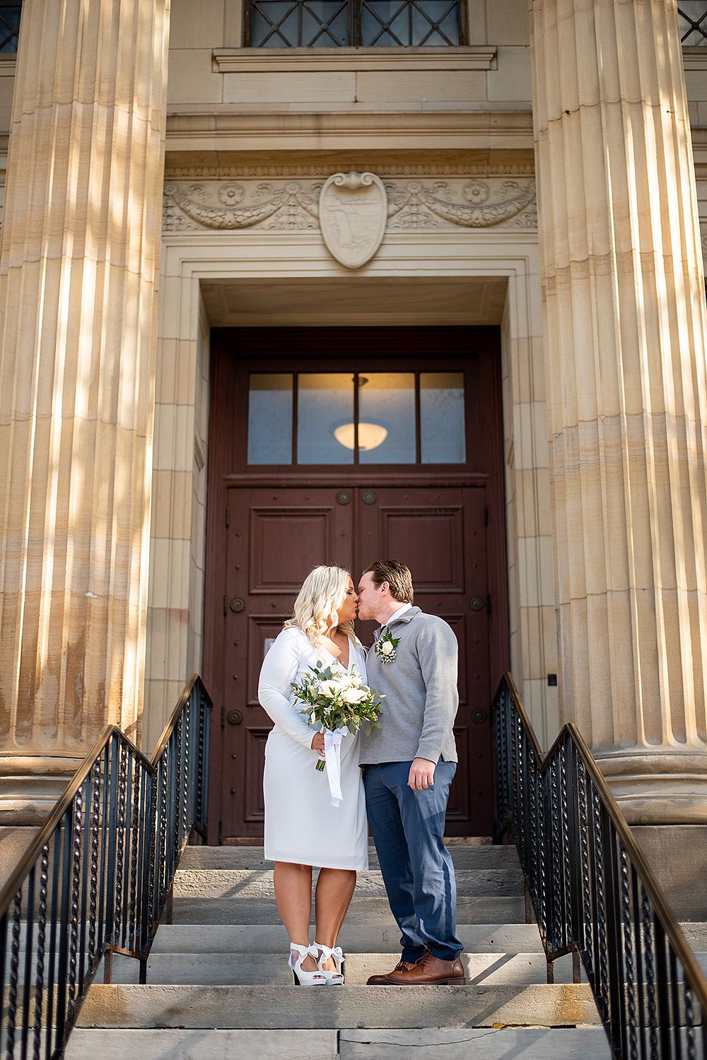 A couple stands on stone steps at a historic building entrance with ornate columns and a brown wooden door.