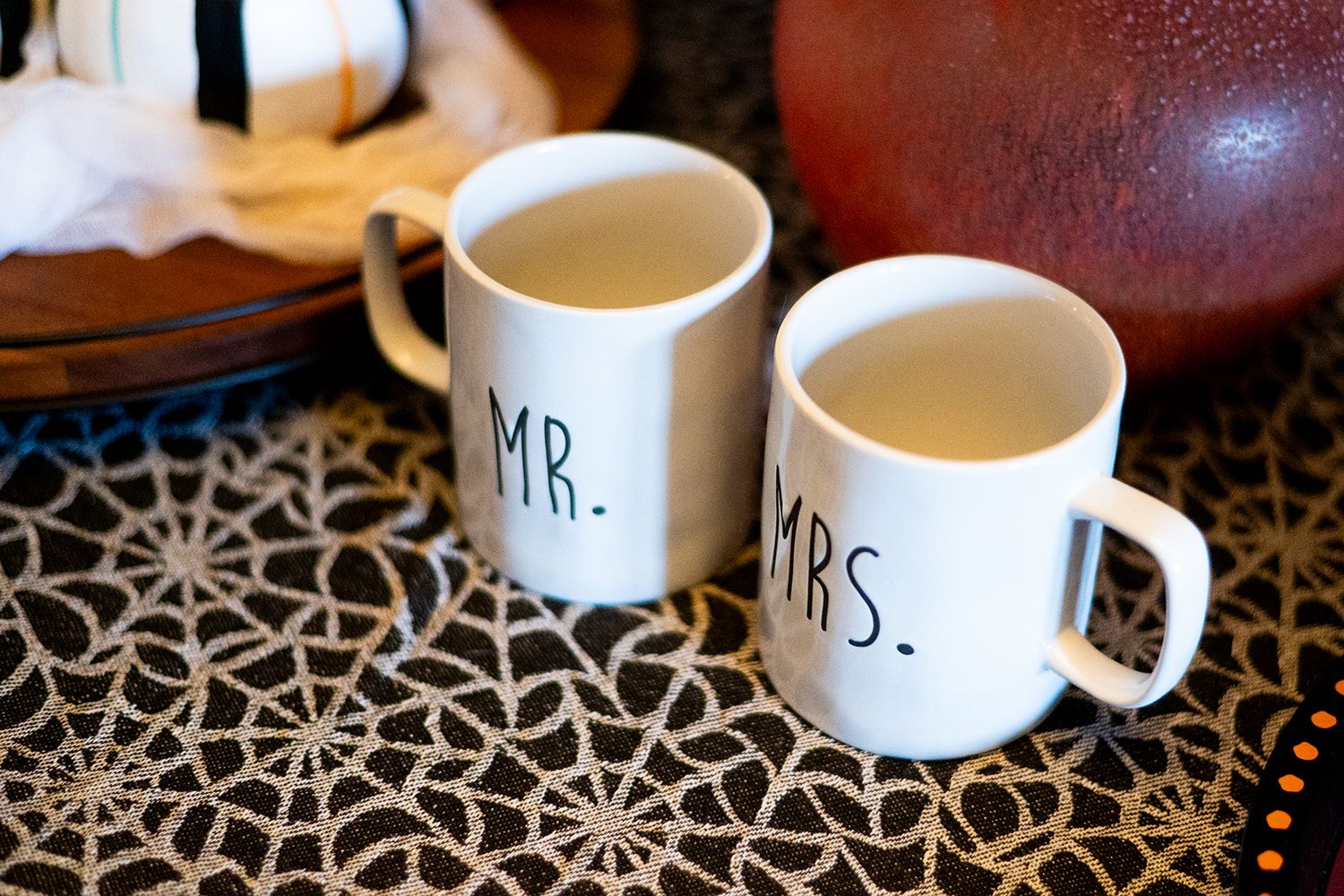 Two matching white ceramic coffee mugs labeled Mr and Mrs sit on a patterned tablecloth with milk and coffee visible.