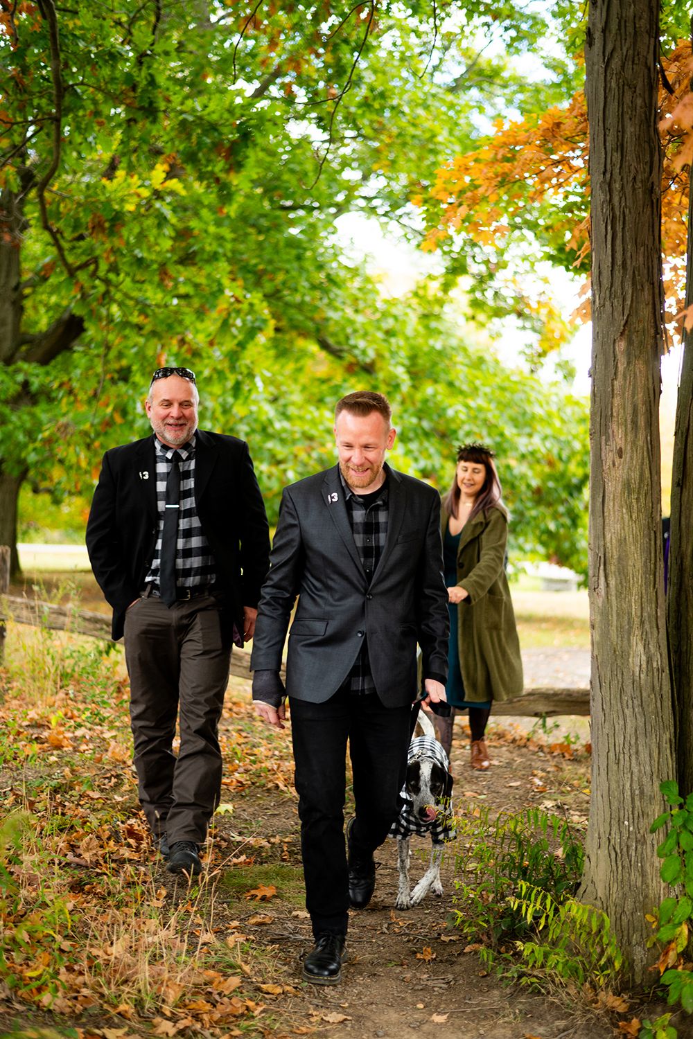 Three people walking down an autumn path through trees with fallen leaves in a scenic park setting.