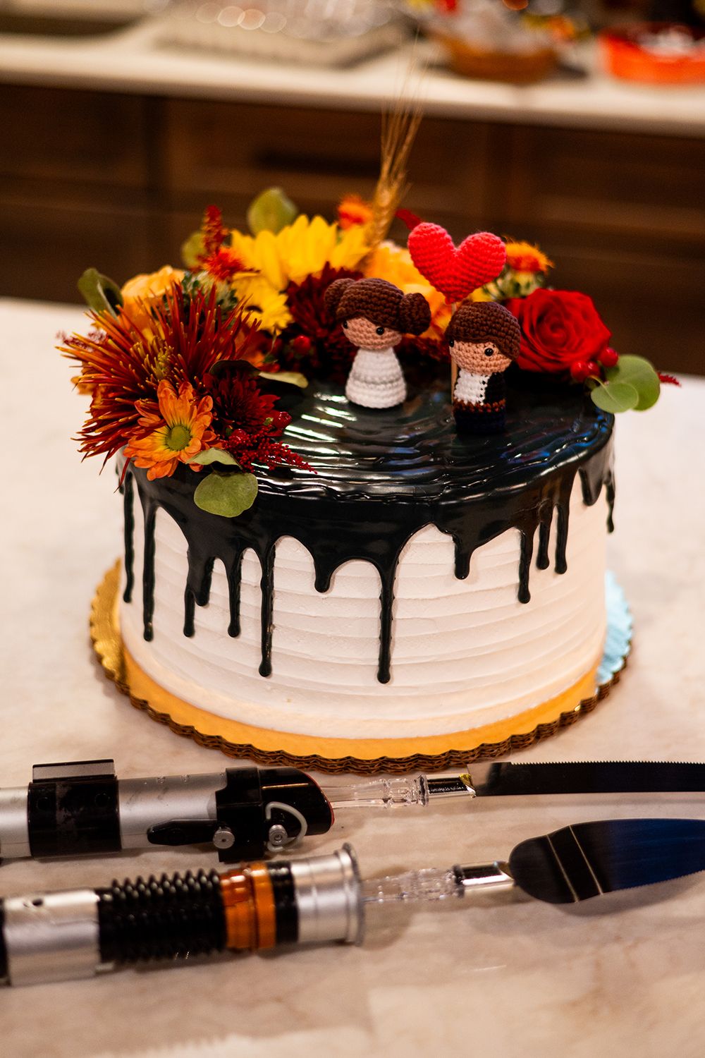 White frosted cake with black chocolate drip, decorated with autumn flowers and a red heart on top next to tattoo equipment.
