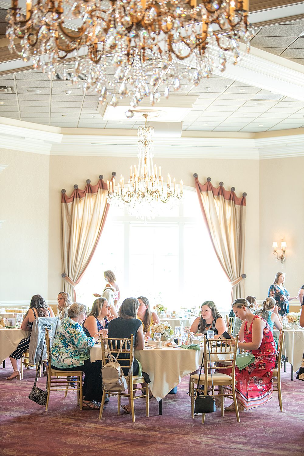 Elegant dining room with crystal chandeliers and ornate drapes features guests seated at round tables for a formal event.