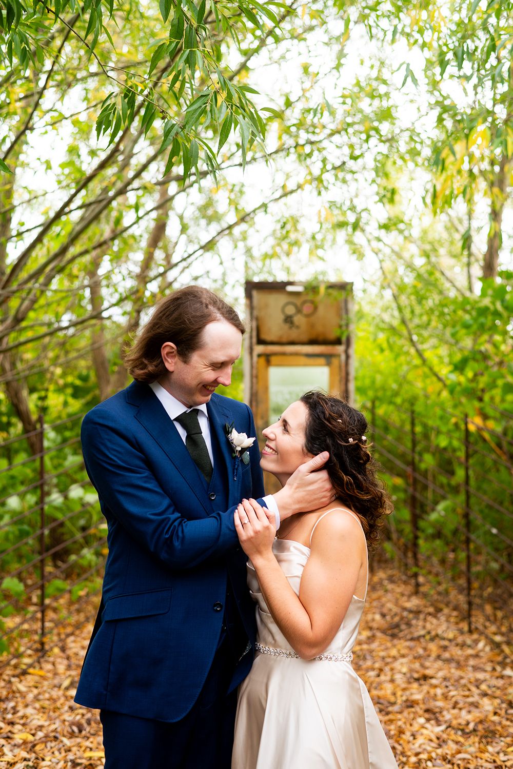Romantic moment captured between newlyweds under green trees with autumn leaves on the ground.