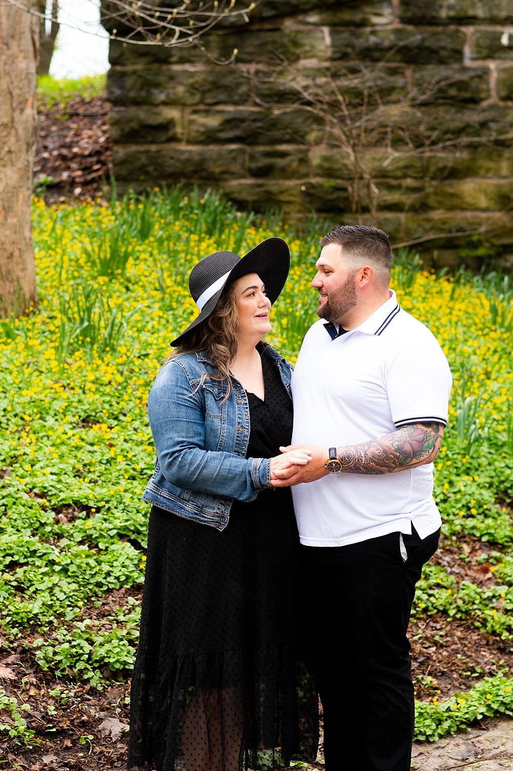 Couple embracing in a field of yellow wildflowers near a stone wall, with her wearing a black hat and denim jacket.